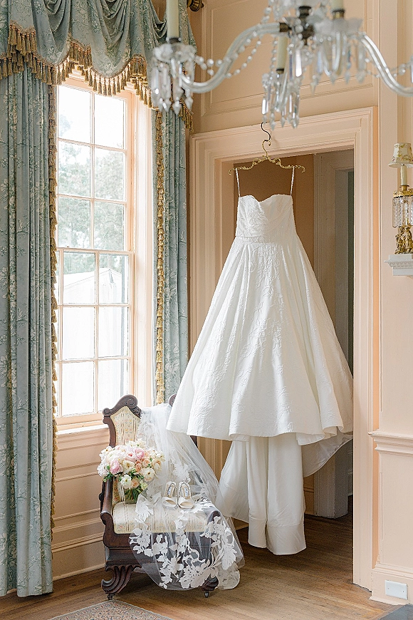 Wedding dress hanging by a tall window, strapless wedding dress with floral embroidery, lace veil, shoes, and bouquet in a paneled room.