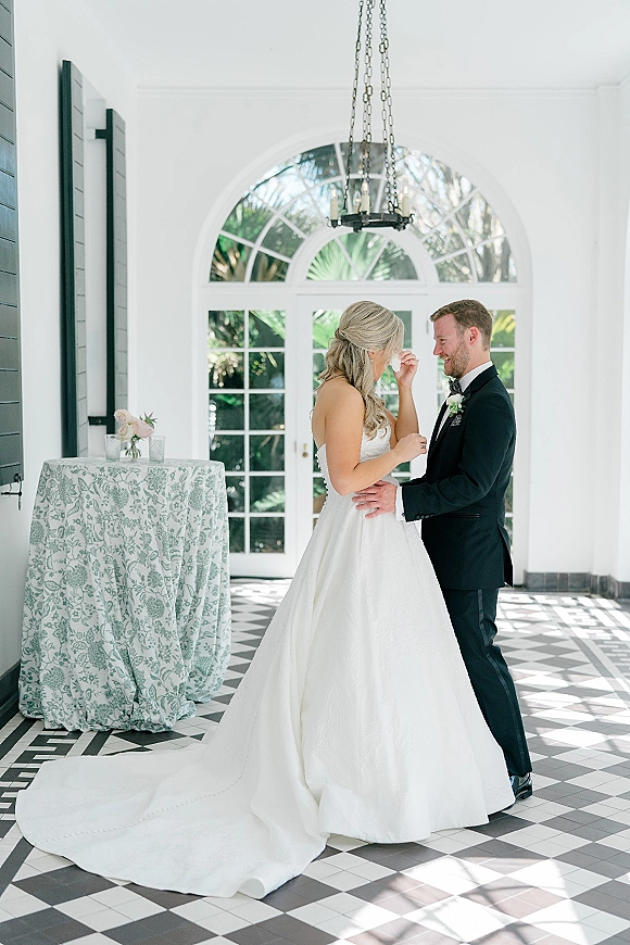 First look moment as bride wipes tears and holds hands with groom in black tux beneath chandelier by arched glass doors