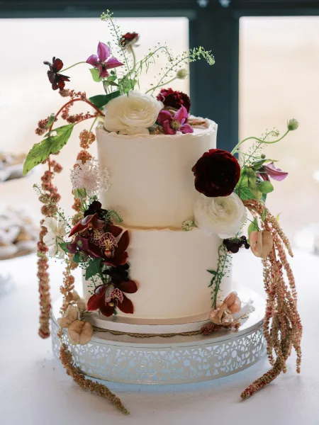 Wedding cake with white buttercream on a stand, topped with cascading ranunculus, orchids, burgundy blooms, and greenery by window light