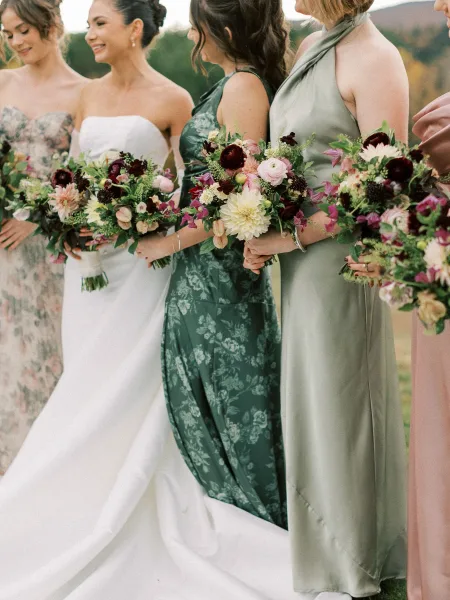 Bridesmaids with bride pose holding bouquets in mixed green and blush dresses, the bride in a gown, in a grassy field with trees and hills