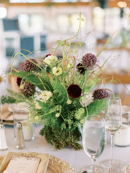 Reception centerpiece with garden style centerpiece florals and greenery on moss, styled with menu card, napkin, gold charger, and glassware
