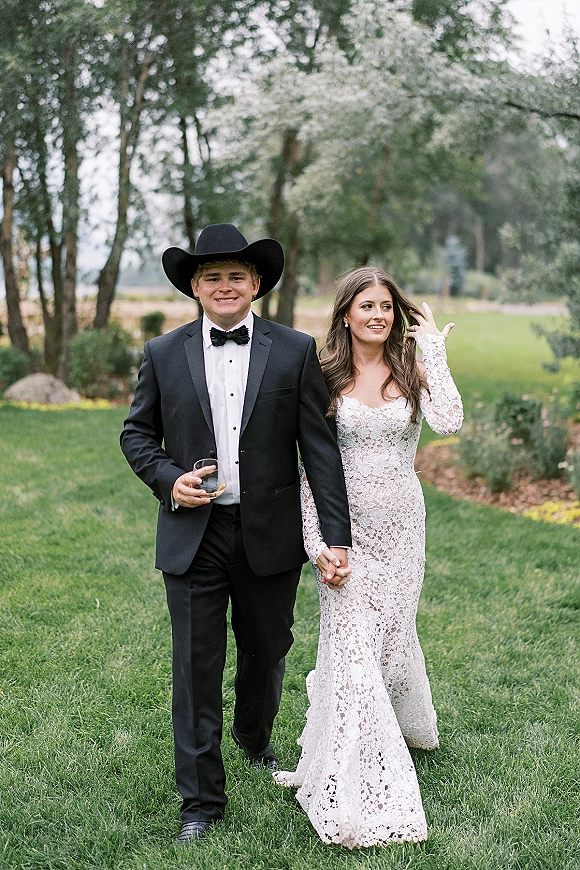 Couple portrait of bride in a lace gown and groom in a cowboy hat holding hands on a green lawn with trees behind them