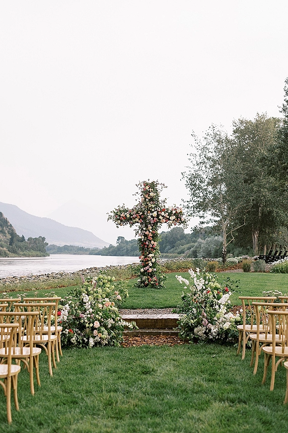 Outdoor ceremony setup with floral cross altar framed by lush greenery, wooden chairs lining an aisle on a riverfront lawn with mountains beyond