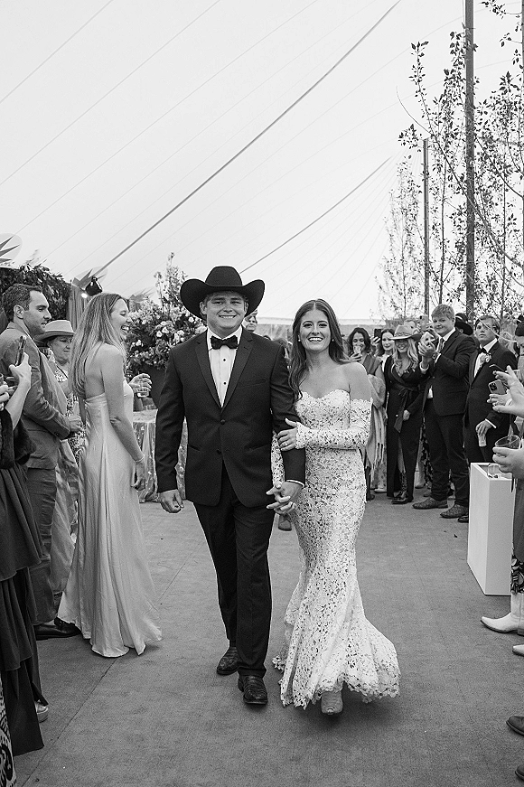Wedding recessional as bride and groom walking aisle under a tent canopy, her lace dress and his cowboy hat, guests cheering with drinks