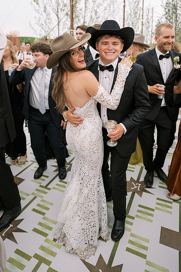 Couple portrait of bride and groom dancing in cowboy hats, her lace off-shoulder gown and his black tuxedo, guests and trees behind