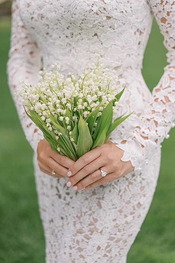 Bridal bouquet close-up of lily of the valley wedding bouquet in lace-sleeved hands with rings and pink manicure against a green lawn