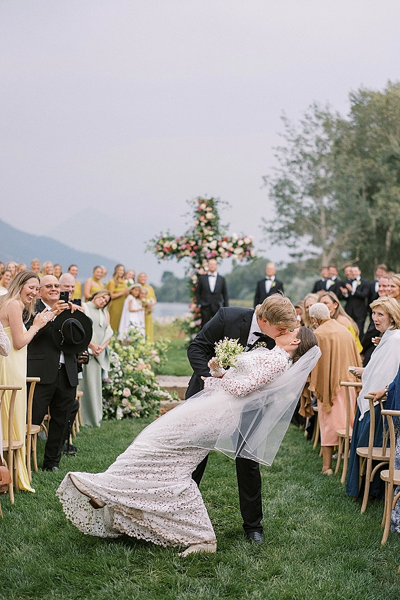 Wedding kiss portrait of bride in lace dress and veil dipping with groom in black tux beneath a floral arch by a mountain lake