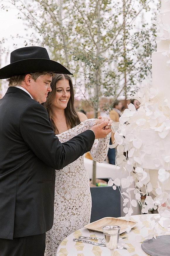 Wedding cake cutting as bride in lace dress and groom share the knife beside a white cake with sugar flowers, cowboy hat, trees behind