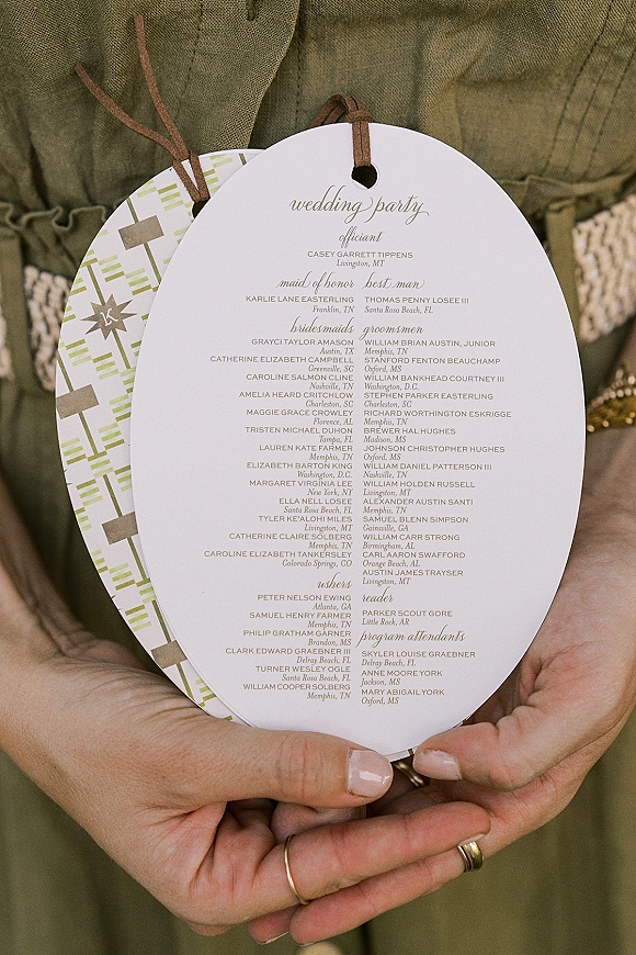 Wedding program wording on die-cut cards tied with a leather cord, held in hands against olive green dress fabric with a gold ring