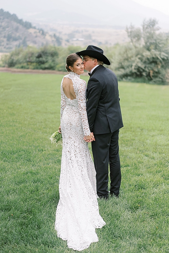 Wedding couple portrait of bride and groom holding hands, groom kissing her cheek as she looks over shoulder on a mountain lawn backdrop