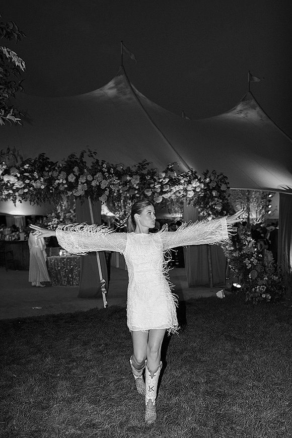 Wedding reception moment of a bride dancing in a short wedding dress with feather fringe sleeves and cowboy boots under a lit tent at night
