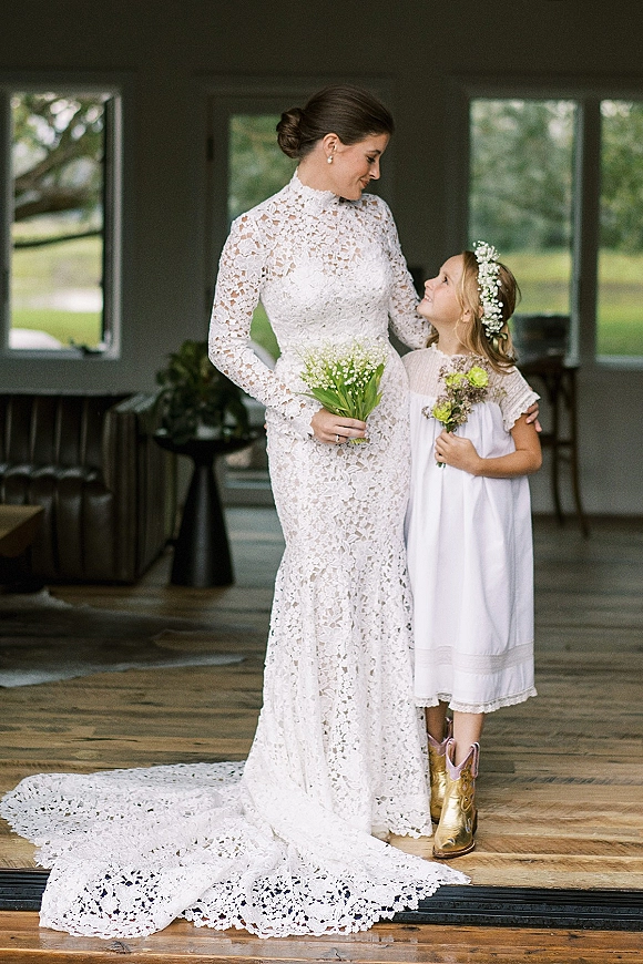Bride and flower girl in lace wedding dress and flower girl with flower crown, holding lily of the valley bouquet on a wooden porch with windows and greenery