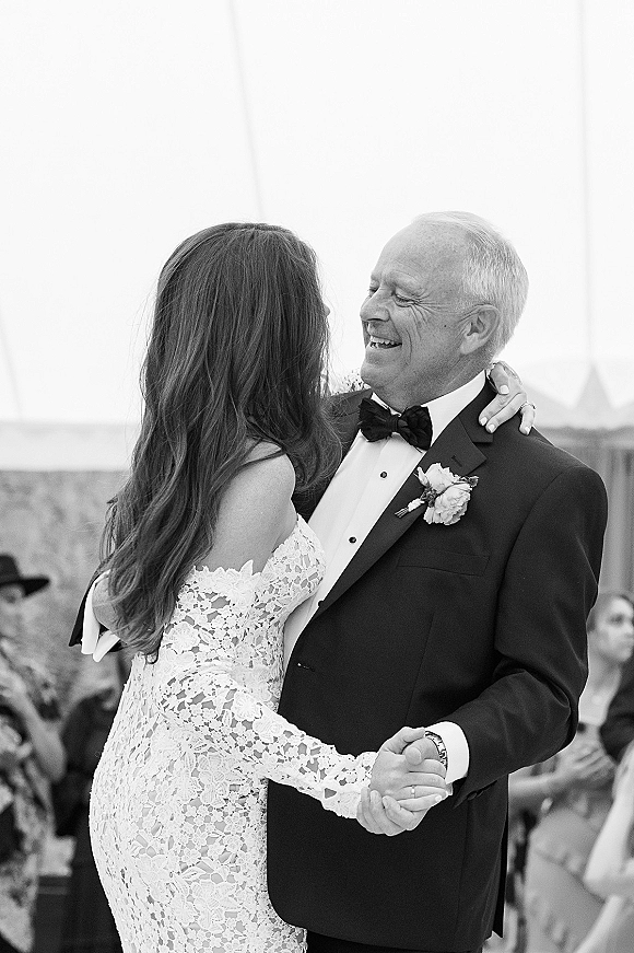 First dance as bride in off-the-shoulder lace dress twirls with father in tuxedo under reception tent, guests watching nearby