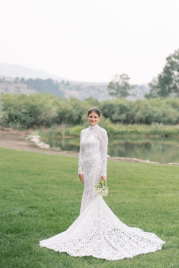 Bridal portrait of a bride in a long sleeve lace wedding dress holding a baby’s breath bouquet by a pond with trees and hills behind