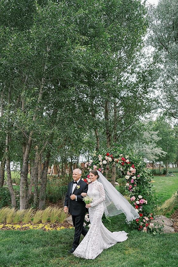 Processional moment as bride walking down aisle in long-sleeve lace gown and veil, holding bouquet beside father on garden lawn under floral arch