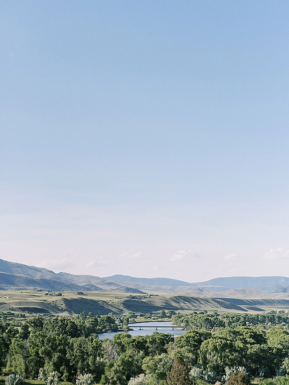 Mountain landscape with a river valley view, rolling hills and tree-lined river bend beneath a wide blue sky and distant ridges