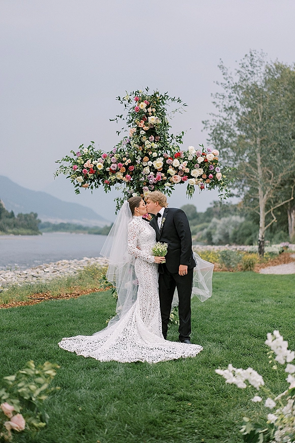 Wedding kiss portrait of bride and groom kissing under a floral cross arch, veil and lace dress by a lakeside with mountains beyond