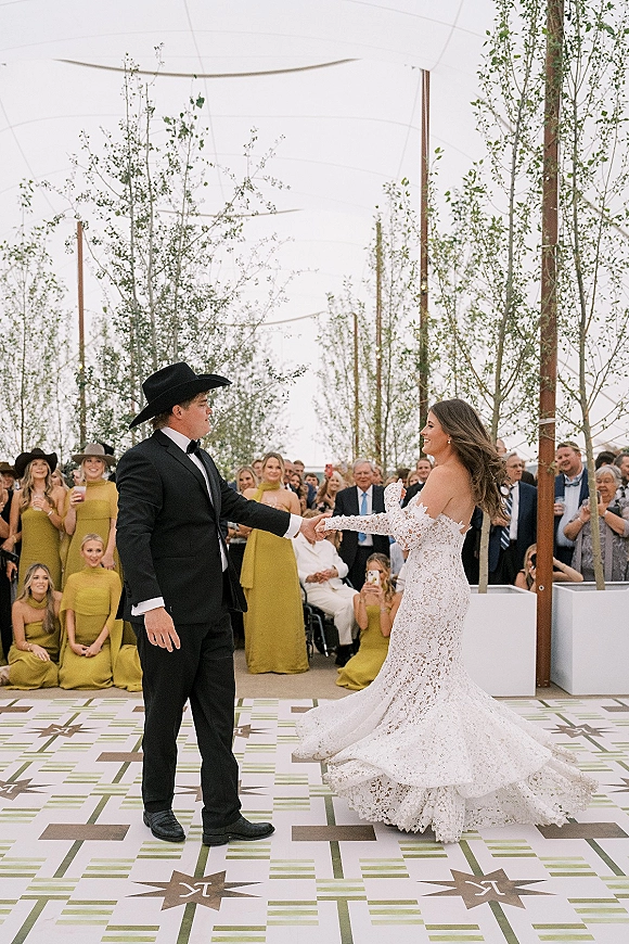 First dance outdoors as bride in lace sleeves twirls while groom in cowboy hat and black tux holds her under tent string lights