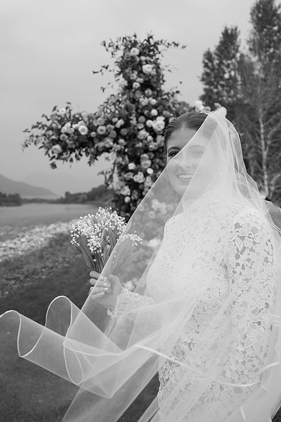 Bridal portrait in black and white of a smiling bride holding a lily of the valley bouquet, veil over face by a mountain lake shoreline