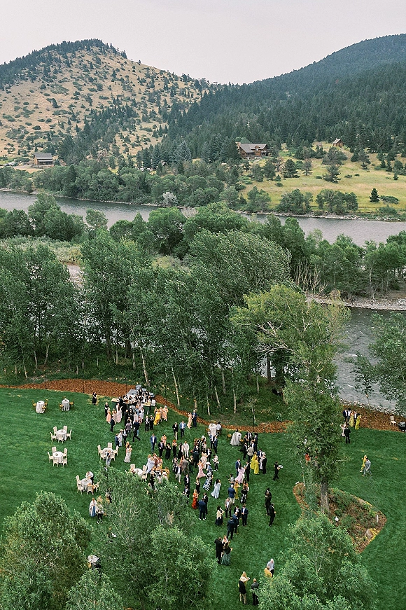 Outdoor wedding reception with wedding cocktail hour setup—cocktail and round tables, chairs, and string lights on a riverside lawn by mountains