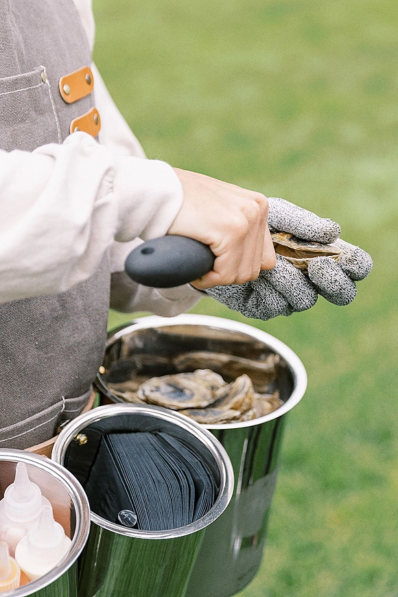 Oyster shucking at a raw oyster bar, hands in cut-resistant glove using an oyster knife, with buckets and squeeze bottles on a grass lawn