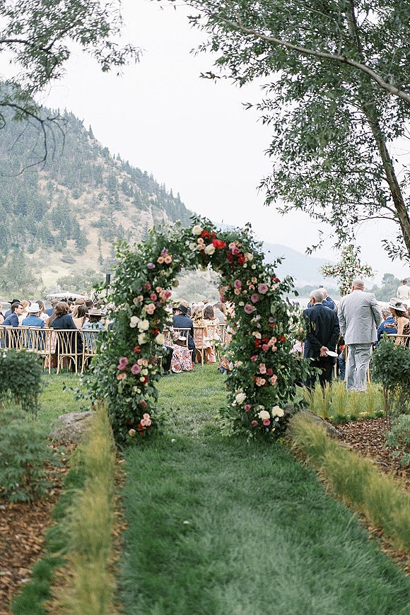 Outdoor ceremony setup with a floral arch lined in roses, garden chairs on a grass aisle, set against a lakeside mountain view