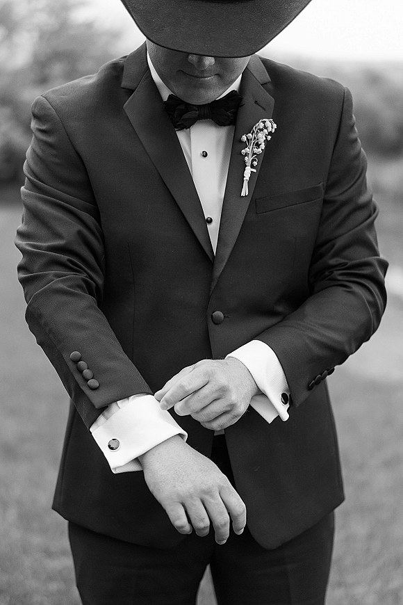 Groom portrait in a tuxedo and bow tie, adjusting cufflinks beside a blurred tree-lined lawn, with boutonniere and fedora hat