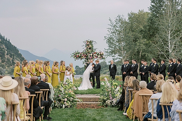 Wedding ceremony with floral cross backdrop as bride and groom stand at the altar, wedding party lined up on a mountain lawn under open sky