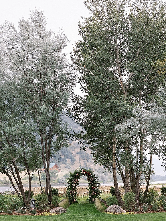 Wedding ceremony arch with roses and greenery swags, framed by wooden chairs and lanterns, set on a lawn by a lake with mountains beyond