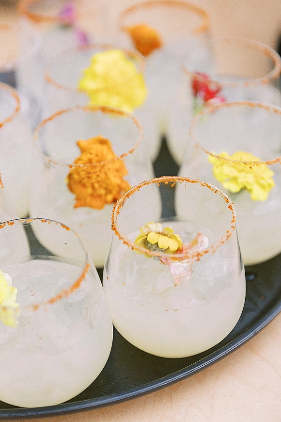 Signature cocktails on a black serving tray with frosted cocktail glasses, ice, chili salt rims, and edible flower accents on a tablecloth