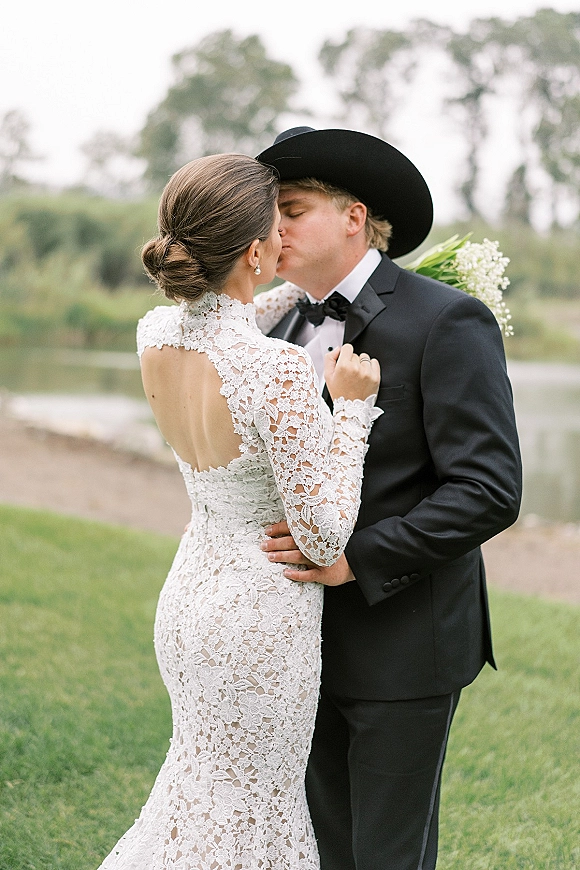 Wedding kiss portrait of bride and groom kissing, groom in cowboy hat and black tuxedo, bride in high-neck lace gown by a lakeside lawn