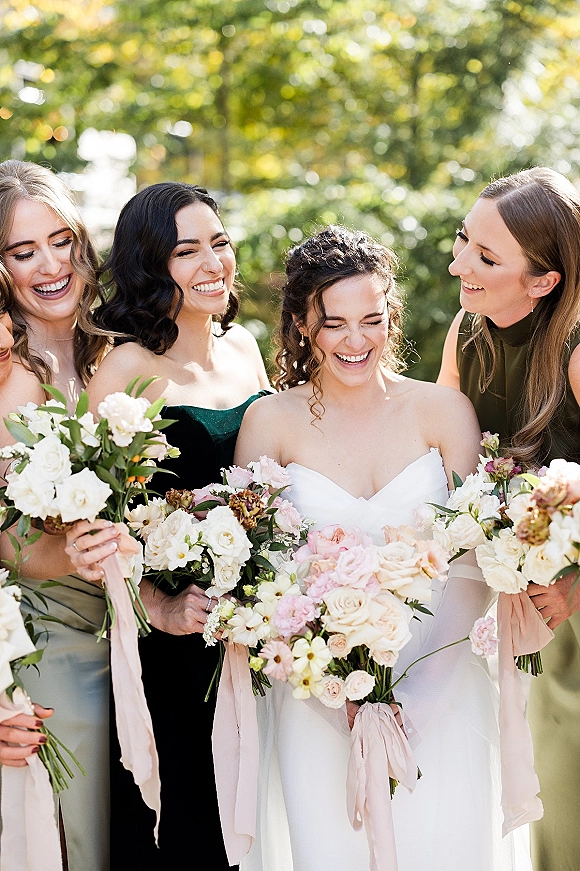 Bridesmaid group photo with bride and bridesmaids laughing, holding white rose and pink bouquets with blush ribbon in a sunlit garden setting