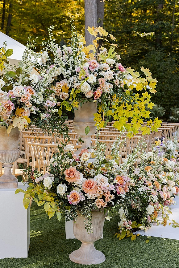 Ceremony floral decor with wedding aisle flowers in stone urns and grounded roses along a white runner leading to a garden tent setting