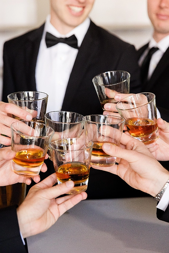 Wedding toast as groomsmen in tuxedos clink whiskey glasses with amber liquor, bow ties and watches visible against an indoor wall