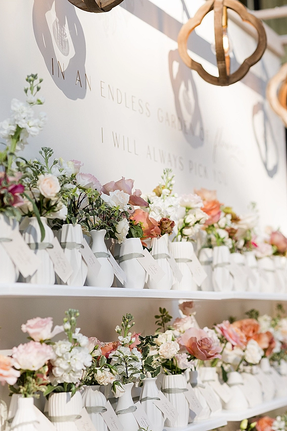 Escort card display with wedding escort cards on white shelves, paired with white bud vases, blush florals, ribbon, and hanging lanterns