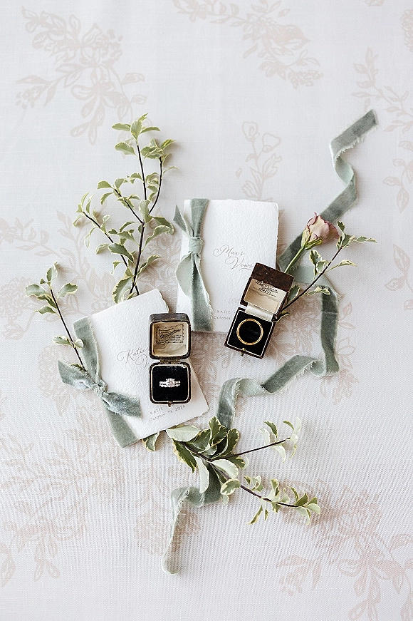 Wedding invitation flat lay with wedding rings flat lay, vow booklets, velvet ring boxes, silk ribbon, greenery and a rosebud on patterned fabric backdrop