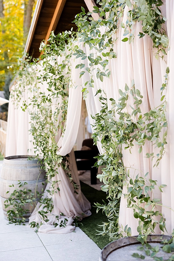 Ceremony backdrop with draped wedding backdrop fabric and greenery garland framed by wooden barrels outside a wood-sided building in daylight