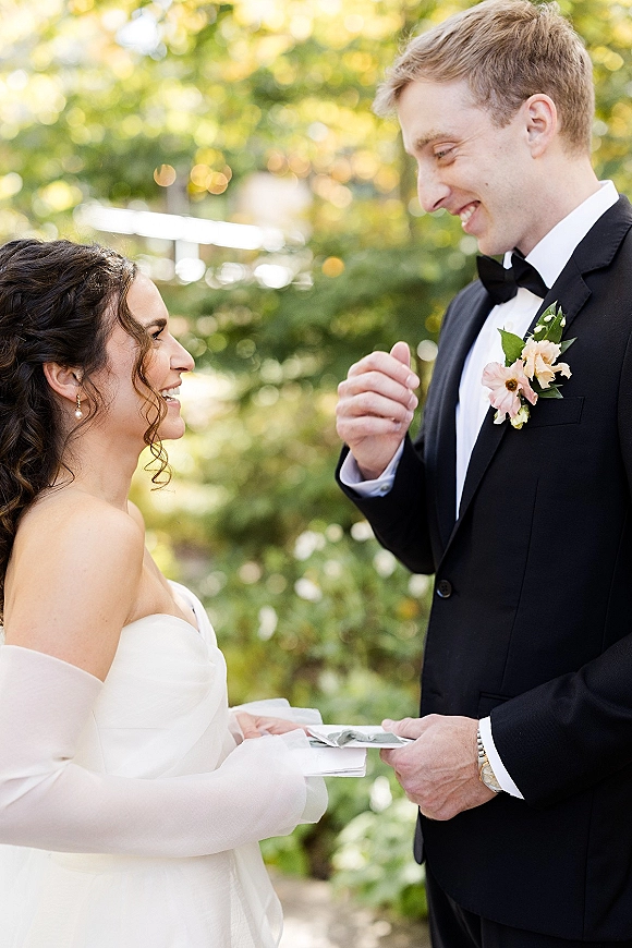 Wedding vows as bride and groom read vow papers, in strapless dress and black tuxedo, surrounded by sunlit garden foliage bokeh