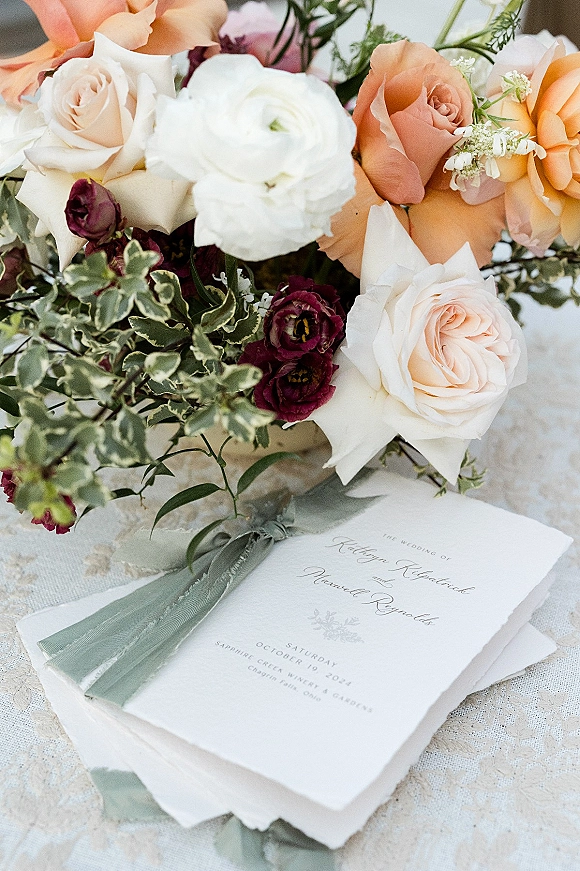 Invitation suite wedding invitation flat lay with deckle-edge card, envelope, and gray silk ribbon beside roses, ranunculus on lace tablecloth