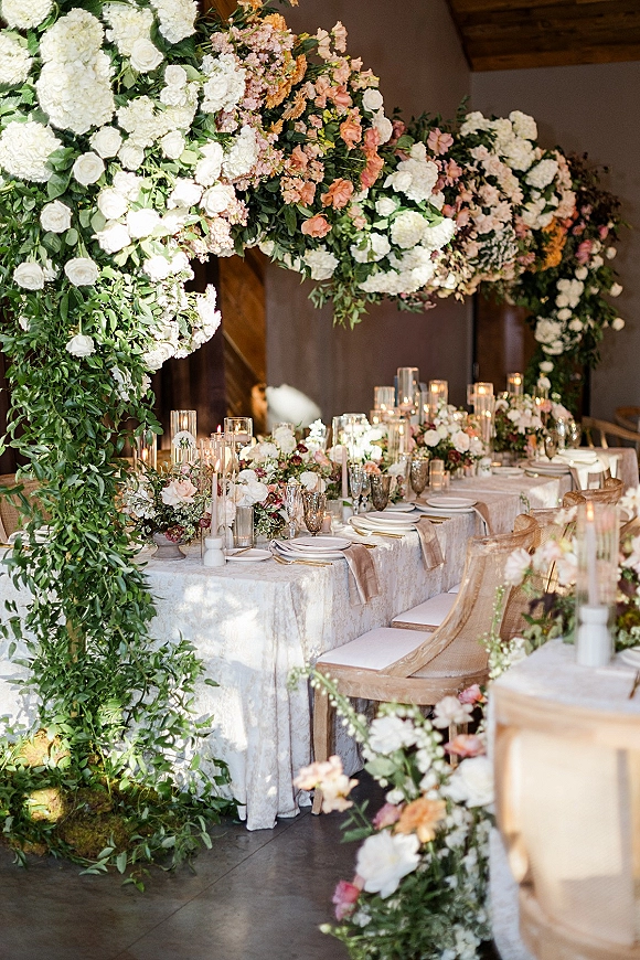 Reception tablescape with wedding head table decor, white and blush florals and candlelit glass vases under a hanging floral canopy indoors