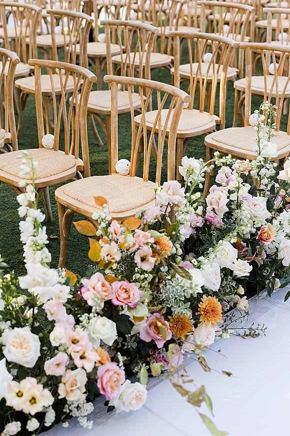 Ceremony seating with wooden chairs lining a white aisle runner, bordered by rose and greenery florals on a grass lawn