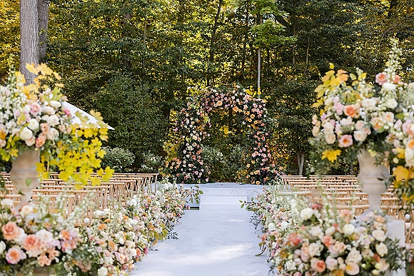 Ceremony setup with a floral arch and aisle flowers along a white runner, wooden chairs facing a platform amid woodland trees