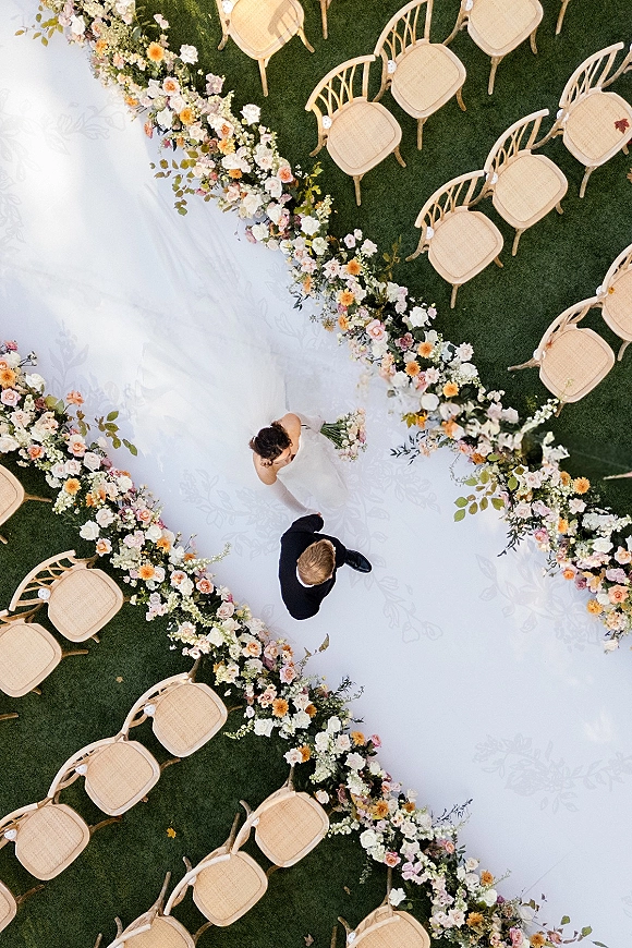 Ceremony aisle decor with a white aisle runner bordered by floral ground arrangements and wooden chairs on a green grass lawn