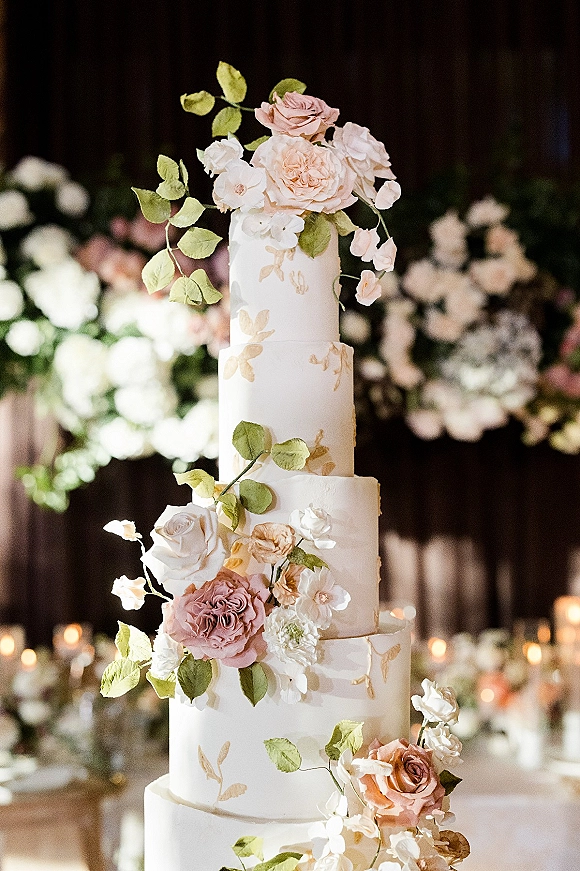 Wedding cake with four tier wedding cake design, white buttercream, sugar roses and greenery cascade, gold leaf details on candlelit table