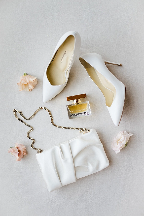 Bridal accessories flat lay featuring wedding shoes flat lay with white pointed-toe heels, bow clutch, gold chain, perfume and blush flowers on gray fabric