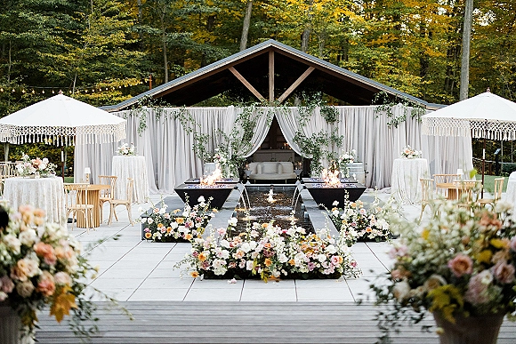 Outdoor reception lounge with wedding lounge seating under white draping, greenery garlands and string lights on a stone patio by woodland trees