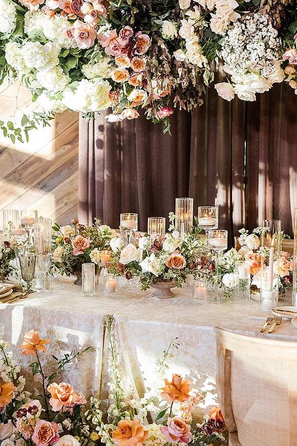 Reception tablescape with overhead floral installation, roses and hydrangea centerpiece, taper and votive candles, crystal glassware, gold flatware, and wood panel backdrop