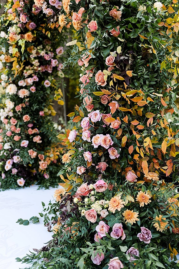 Floral ceremony arch with roses and dahlias, lush greenery, and autumn leaves framed by garden trees and outdoor greenery backdrop