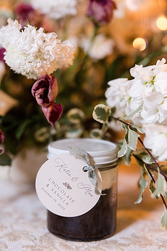 Wedding favor jars with metal lids and personalized round tags tied with ribbon, styled on a lace runner with warm bokeh lights behind
