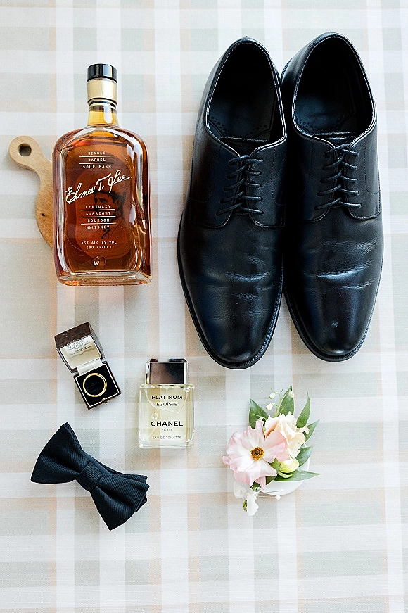 Groom details flatlay with black dress shoes, bourbon whiskey bottle, ring box, cologne, and bow tie on a checkered tablecloth